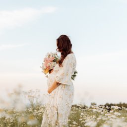 Karolina Cieszyńska fotografia - Kobieta w ciąży w kwiecistej sukience, trzymająca bukiet kwiatów, stoi bokiem na łące pełnej białych margerytek.
