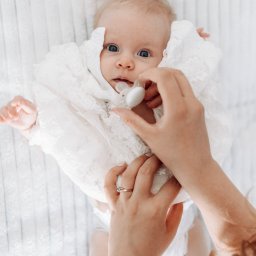 Karolina Cieszyńska fotografia - Niemowlę leżące na białym, teksturowanym tle, ubrane w koronkowy strój, z ręką dorosłego podającą smoczek; widoczne niebieskie oczy dziecka i pierścionek z turkusem na palcu dorosłej osoby.