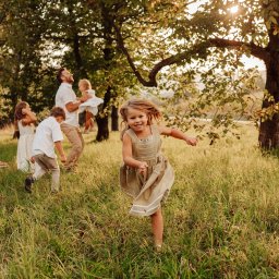 Karolina Cieszyńska fotografia - Uśmiechnięta dziewczynka biegnie przez łąkę w sukience w stylu retro, w tle rodzina z dziećmi w jasnych ubraniach i zachodzące słońce przebijające przez liście drzew