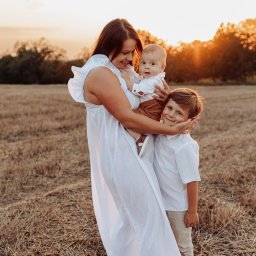 Karolina Cieszyńska fotografia - Uśmiechnięta matka w białej sukience trzymająca niemowlę i obejmowana przez starszego syna na tle pola o zachodzie słońca.