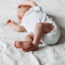 Karolina Cieszyńska fotografia - Noworodek w białym body leży na białej, teksturowanej kołderce, z bliska widoczne małe stopy z delikatnymi palcami.
