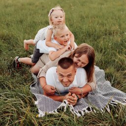 Karolina Cieszyńska fotografia - Rodzina leżąca na kocu w trawie: tata z uśmiechem patrzy w obiektyw, mama śmieje się obok, a na nich leżą dwójka dzieci - syn i córka z kokardą we włosach.