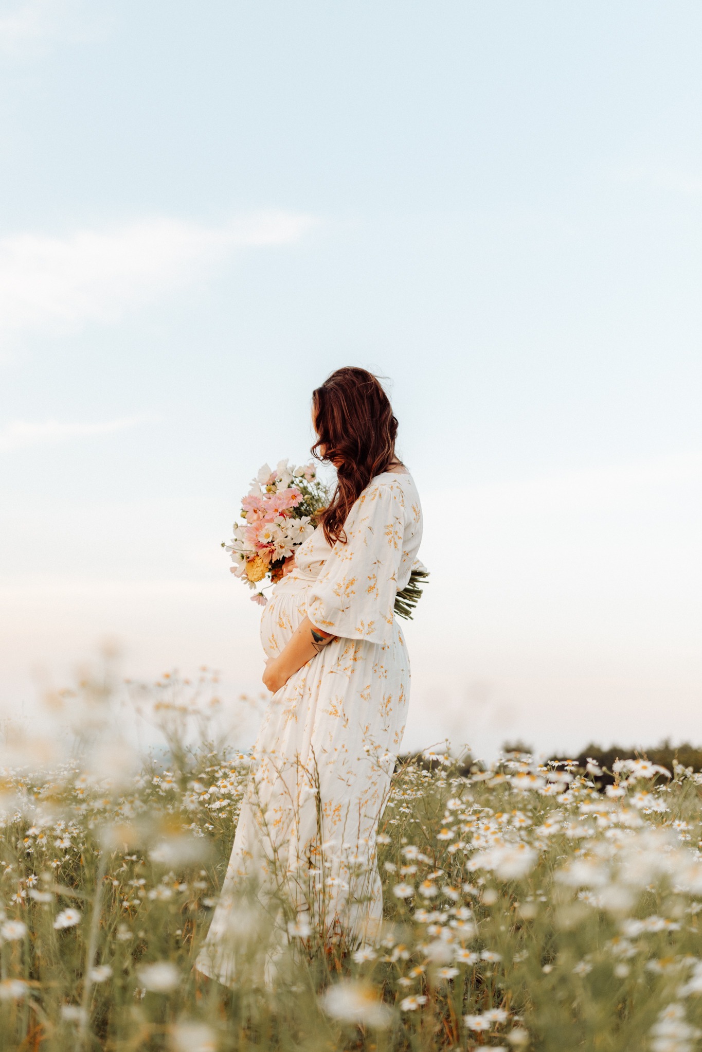 Kobieta w ciąży w kwiecistej sukience, trzymająca bukiet kwiatów, stoi bokiem na łące pełnej białych margerytek.
