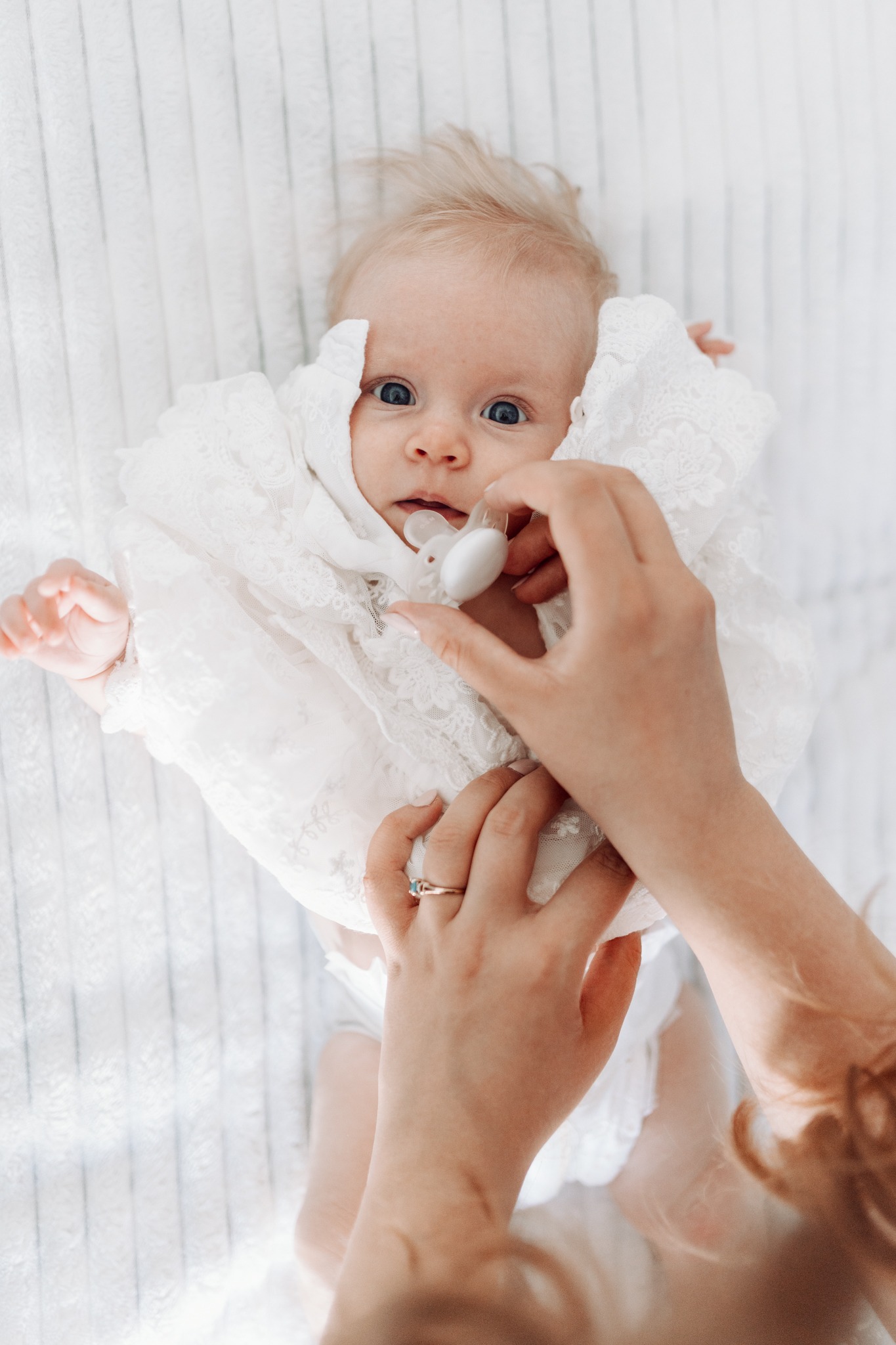 Niemowlę leżące na białym, teksturowanym tle, ubrane w koronkowy strój, z ręką dorosłego podającą smoczek; widoczne niebieskie oczy dziecka i pierścionek z turkusem na palcu dorosłej osoby.