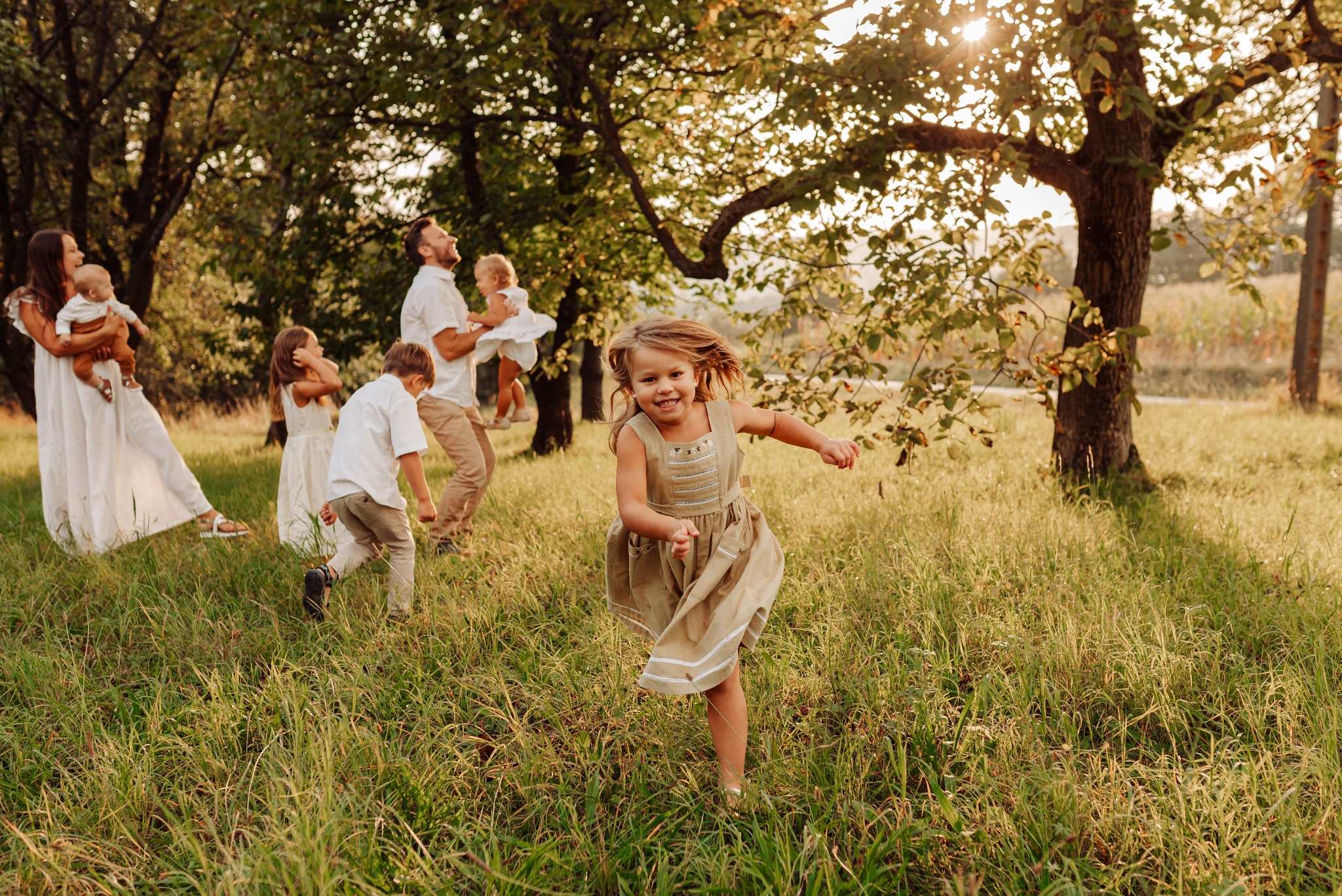 Uśmiechnięta dziewczynka biegnie przez łąkę w sukience w stylu retro, w tle rodzina z dziećmi w jasnych ubraniach i zachodzące słońce przebijające przez liście drzew