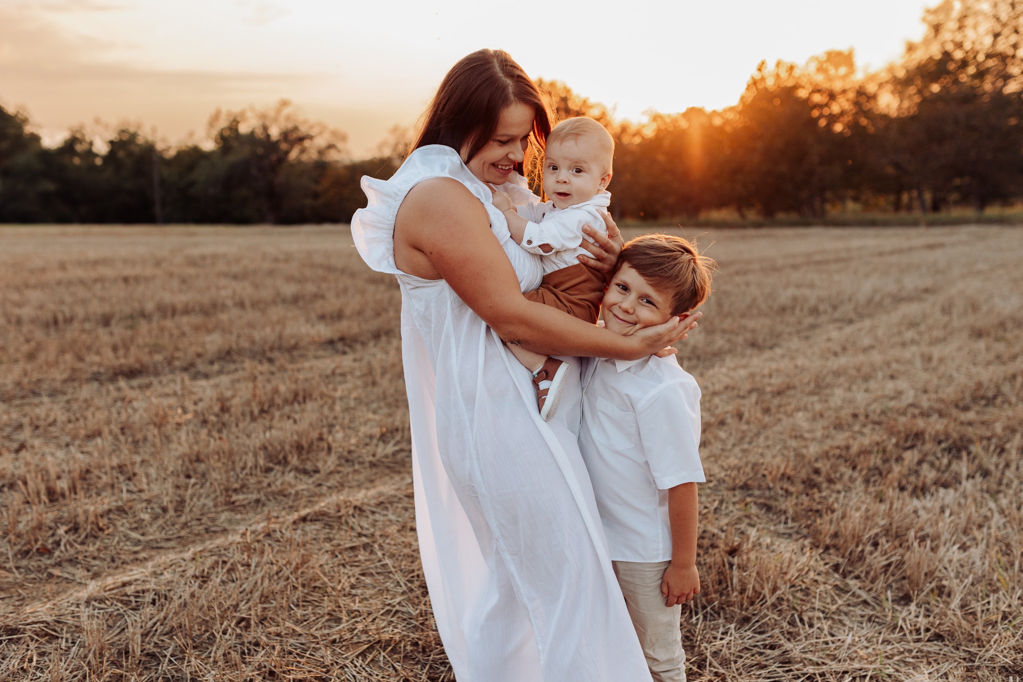 Uśmiechnięta matka w białej sukience trzymająca niemowlę i obejmowana przez starszego syna na tle pola o zachodzie słońca.
