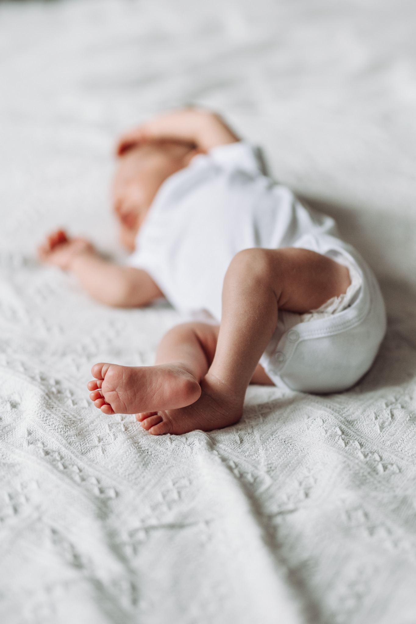 Noworodek w białym body leży na białej, teksturowanej kołderce, z bliska widoczne małe stopy z delikatnymi palcami.