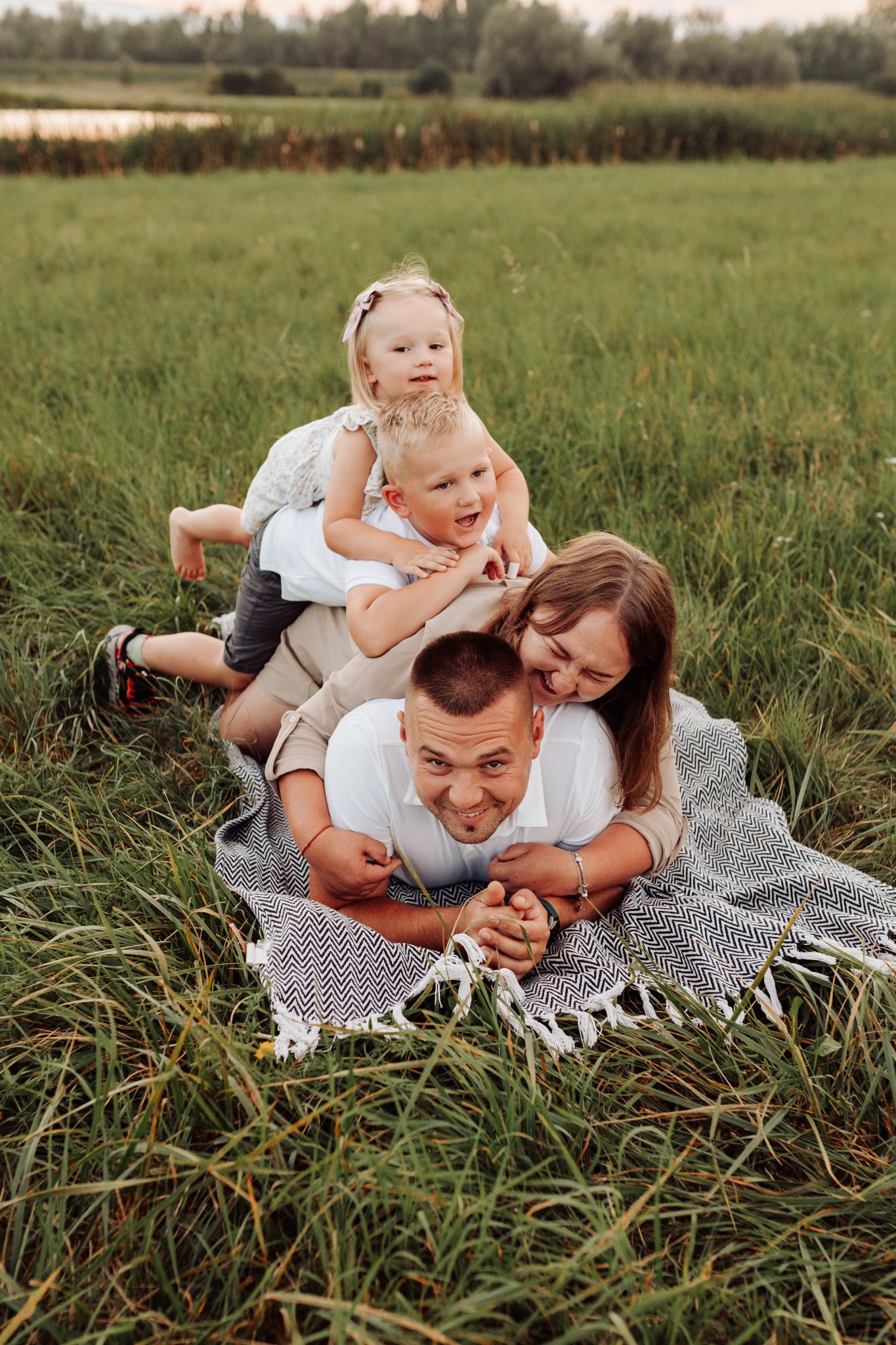 Rodzina leżąca na kocu w trawie: tata z uśmiechem patrzy w obiektyw, mama śmieje się obok, a na nich leżą dwójka dzieci - syn i córka z kokardą we włosach.