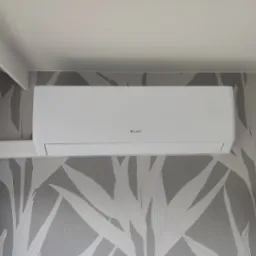 Biały klimatyzator marki Gree zamontowany na ścianie z tapetą w delikatne, szaro-białe liście. Widoczny fragment sufitu i telewizora.