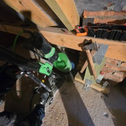 ClimaTex - Wiertnica Eibenstock z zielonym zbiornikiem na wodę, leżąca na poddaszu, obok młotek i elementy konstrukcji drewnianej. Widoczna stopa osoby obsługującej urządzenie.