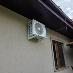 ClimaTex - Biała jednostka zewnętrzna klimatyzacji marki Gree zamontowana na elewacji budynku z brązowym dachem i oknem. Widoczne mocowania i fragment przewodu.