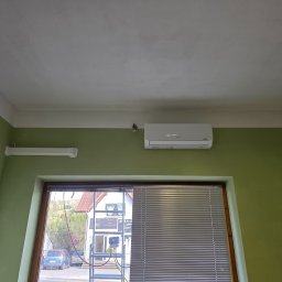 ClimaTex - Biały klimatyzator Gree zamontowany na zielonej ścianie nad oknem z żaluzjami. Na zewnątrz drabina z urządzeniem do serwisu klimatyzacji.