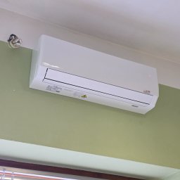 ClimaTex - Biała klimatyzacja ścienna na zielonej ścianie, widoczny fragment okna z żaluzjami. Instalacja elektryczna obok urządzenia.