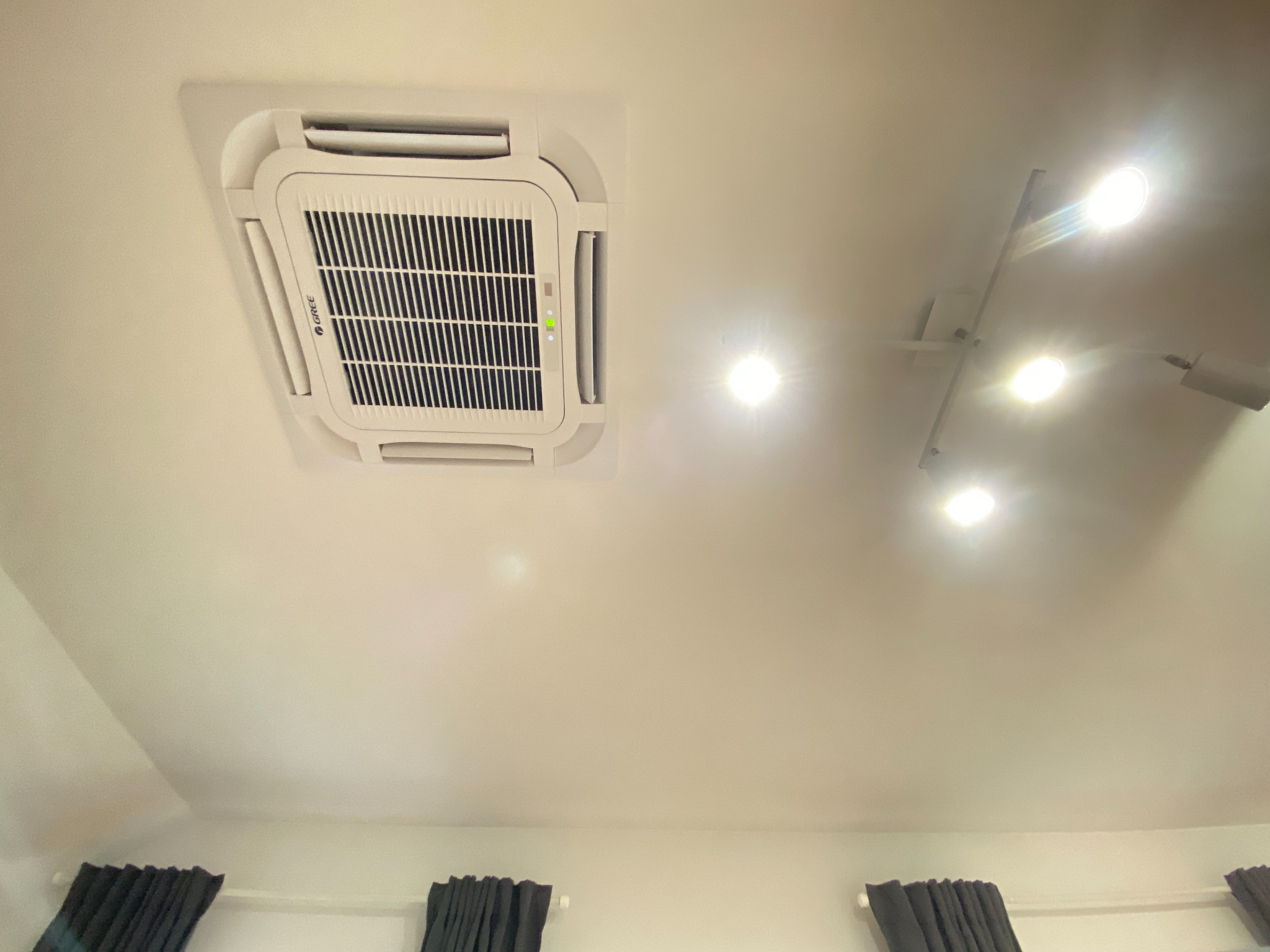 Kasetonowa jednostka klimatyzacyjna marki Gree zamontowana w suficie podwieszanym, widoczna z dołu, z włączonymi diodami sygnalizacyjnymi, oświetlona przez reflektory punktowe.