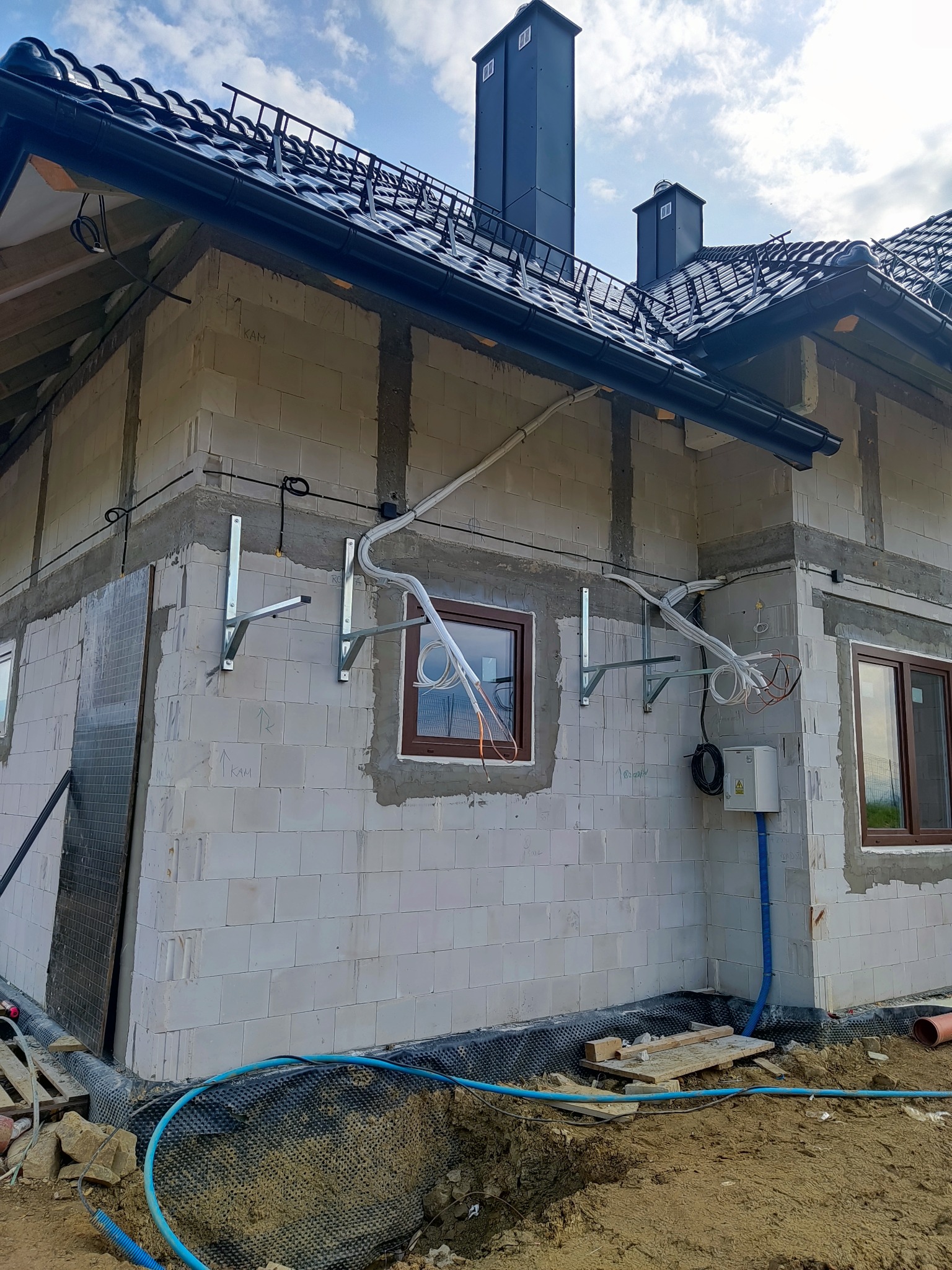 Surowa ściana budynku w trakcie budowy z przygotowanymi uchwytami i przewodami do montażu instalacji zewnętrznej, widoczna skrzynka elektryczna i częściowo wykopane fundamenty.