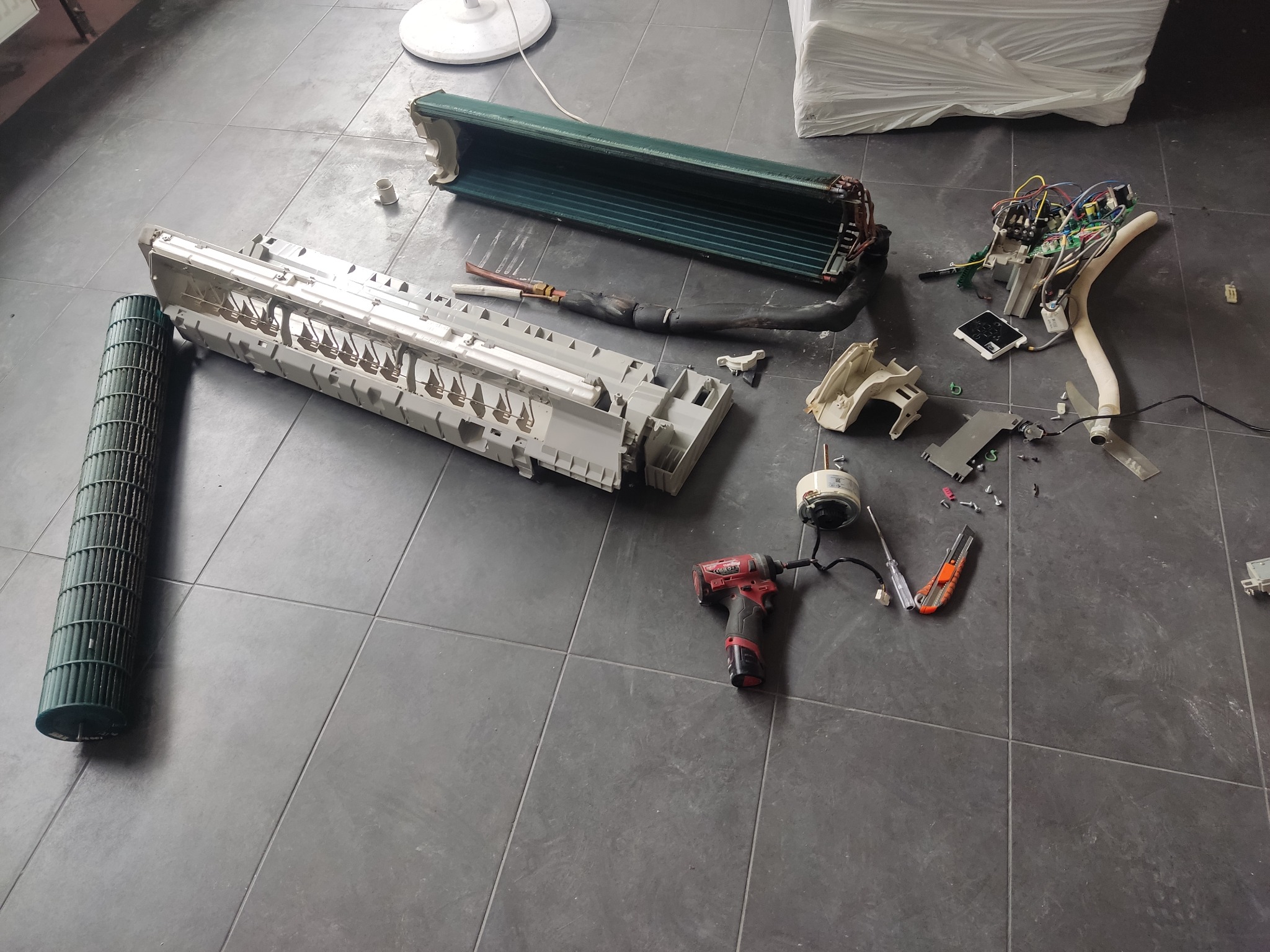 Rozłożony na podłodze klimatyzator podczas serwisu, widoczne elementy wewnętrzne, obudowa, wentylator bębnowy, narzędzia: wkrętarka udarowa i nóż monterski.
