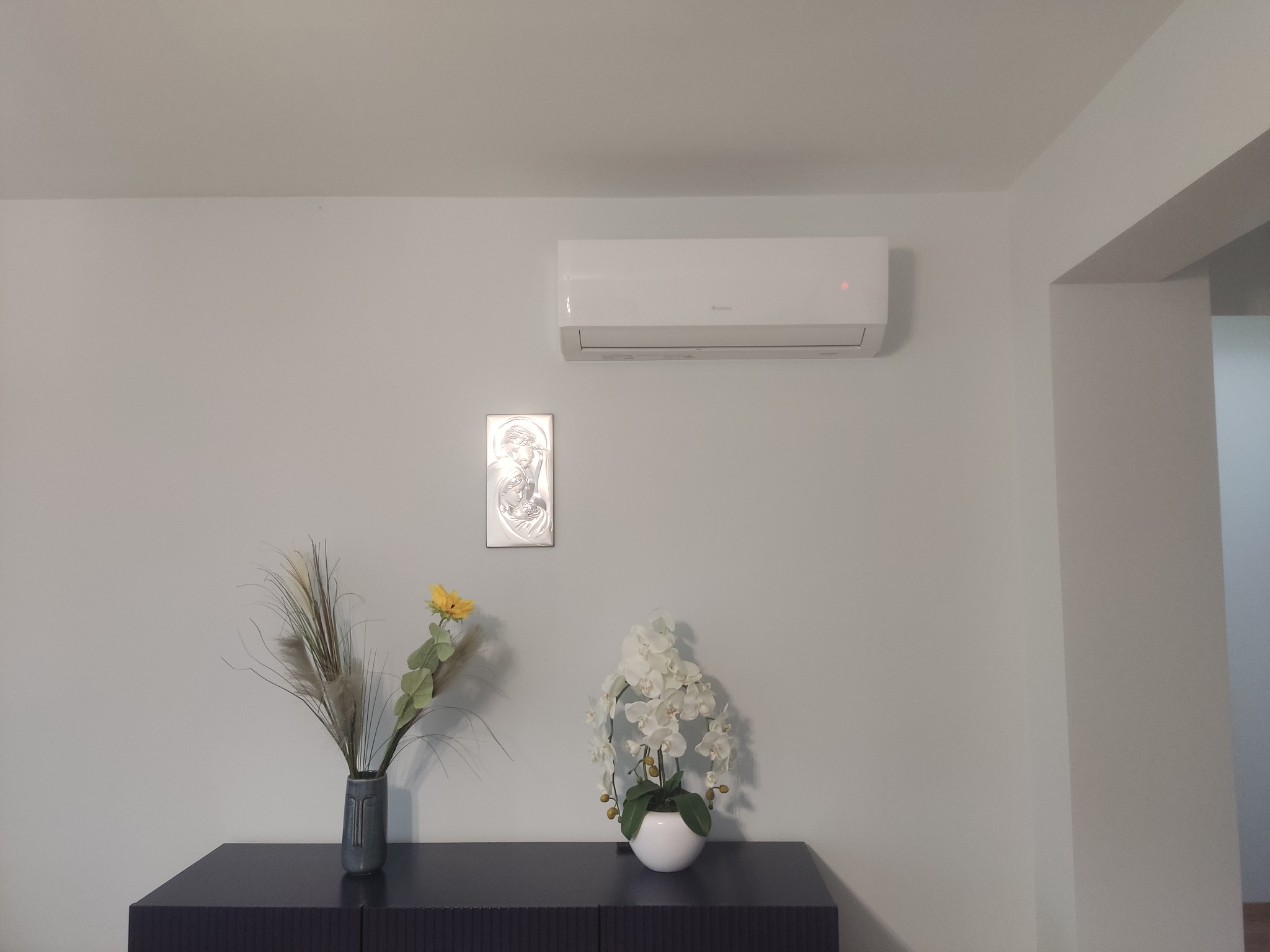 Biały klimatyzator ścienny marki Gree zamontowany w minimalistycznym wnętrzu nad ciemnogranatową komodą z dekoracjami: wazonem z suszem i kompozycją z białych kwiatów, obok srebrny obrazek.