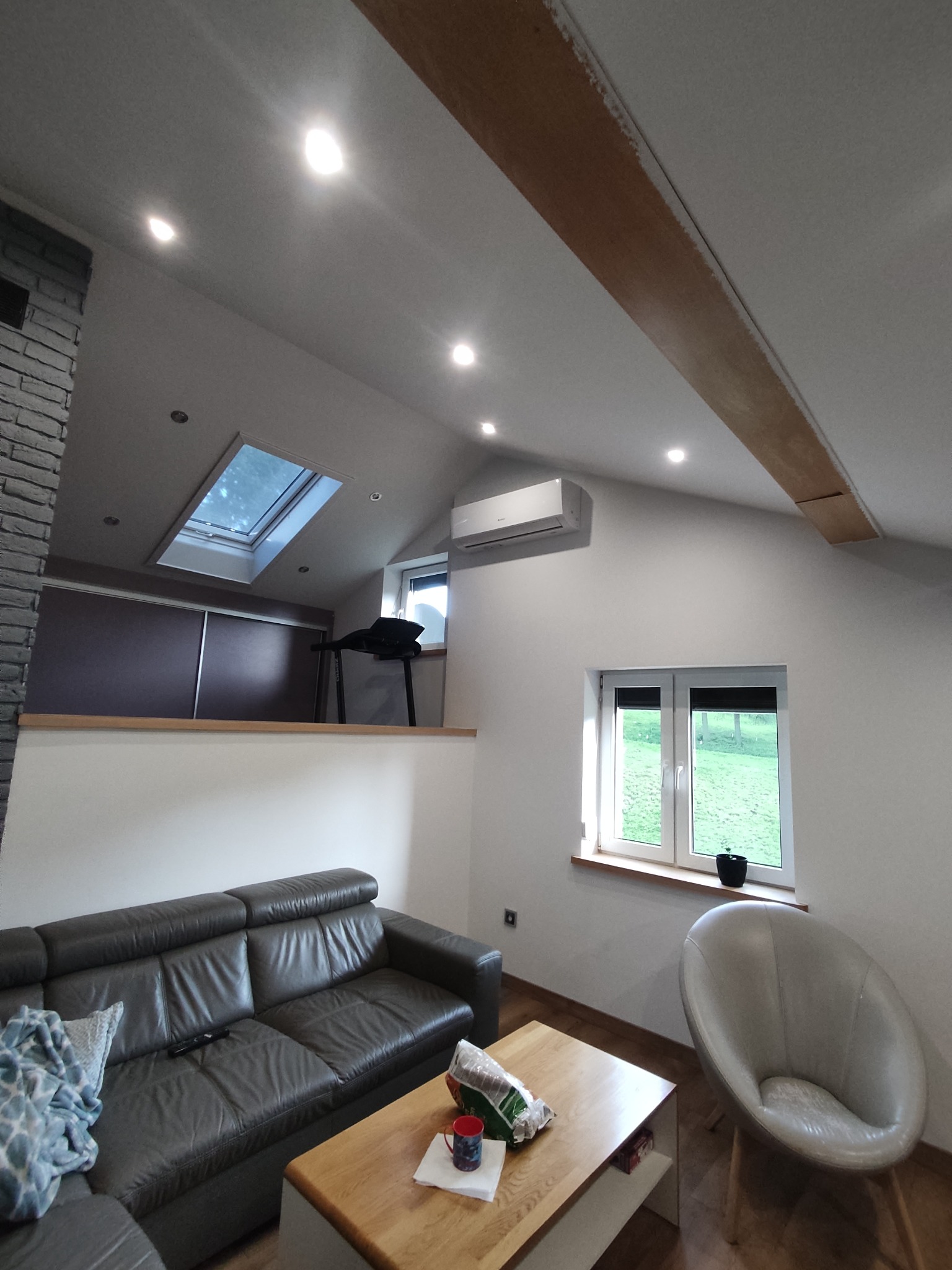 Wnętrze poddasza z klimatyzatorem ściennym, widocznym nad oknem, oraz oknem dachowym, z drewnianymi belkami stropowymi, szarą sofą, drewnianym stolikiem kawowym i designerskim fotelem.