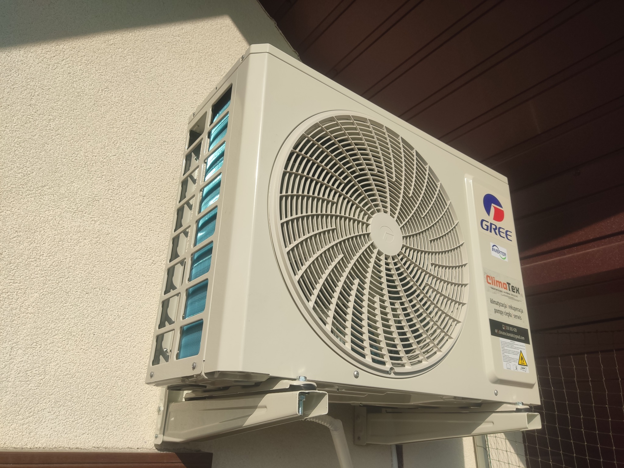 Zewnętrzna jednostka klimatyzacji marki Gree zamontowana na elewacji budynku, widoczna z dołu pod kątem, z logo i naklejką informacyjną firmy ClimaTex.