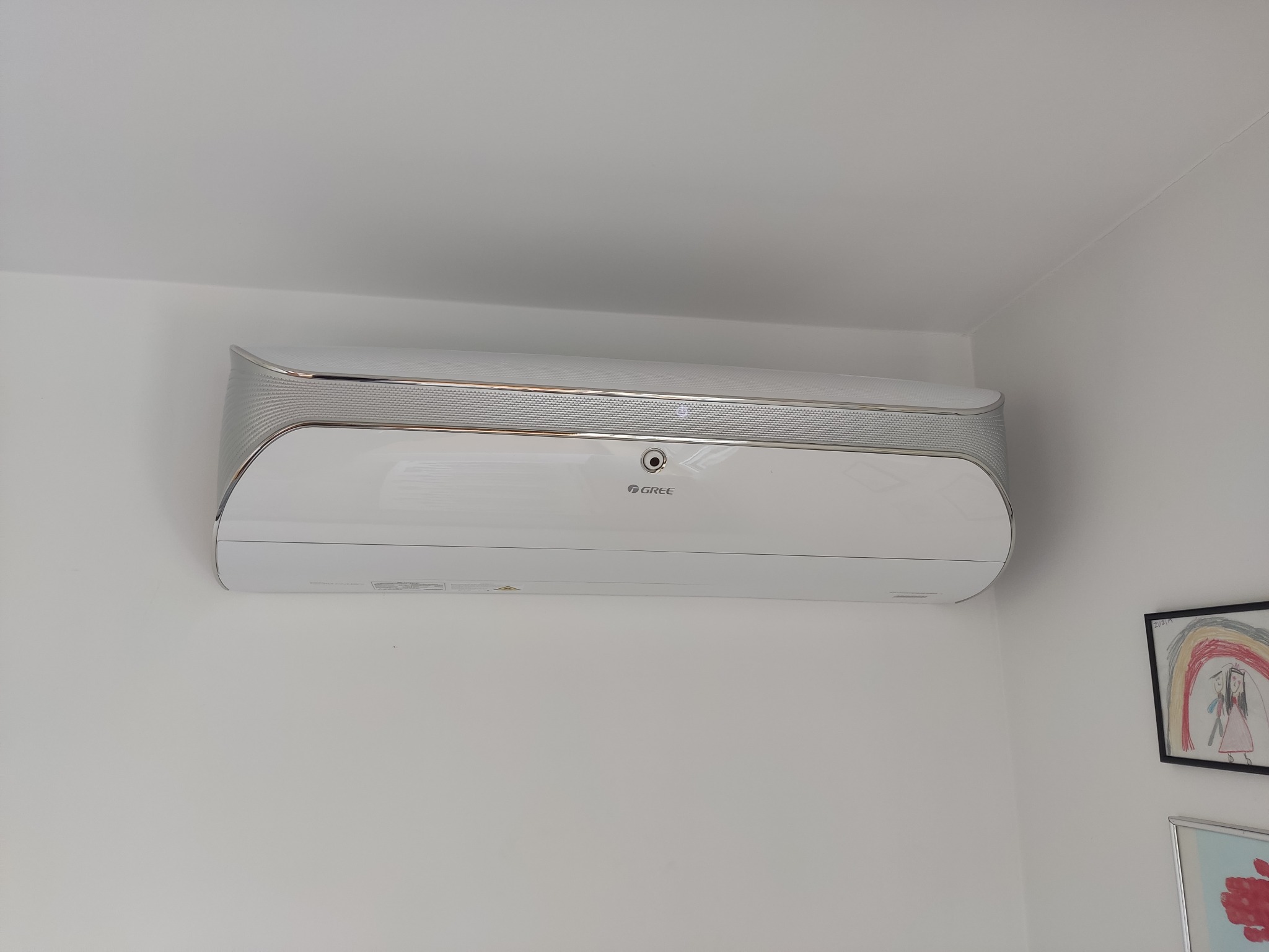 Nowoczesny, biały klimatyzator marki Gree zamontowany na białej ścianie w pomieszczeniu mieszkalnym, widoczne rysunki dziecięce w ramkach w tle.