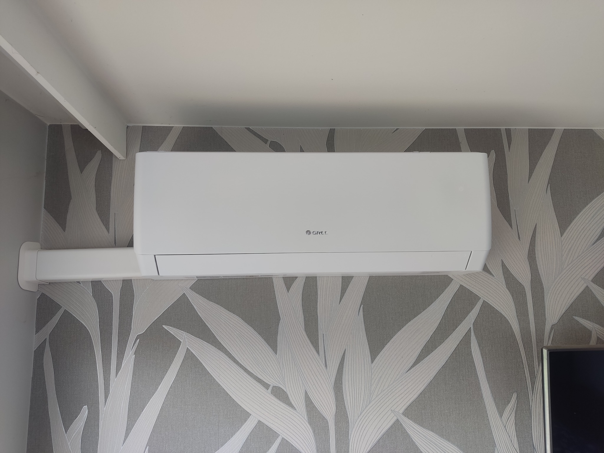 Biały klimatyzator marki Gree zamontowany na ścianie z tapetą w delikatne, szaro-białe liście. Widoczny fragment sufitu i telewizora.
