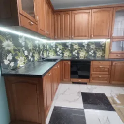 Klasyczna kuchnia z drewnianymi frontami, czarnym blatem i tapetą w kwiaty. Podświetlenie LED pod szafkami górnymi. Na podłodze biało-czarna szachownica. Częściowo zabezpieczona podłoga kartonem.