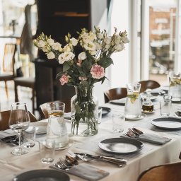 RAFAŁ KOZIORZEMSKI - Elegancko nakryty stół z ciemnymi talerzami, srebrnymi sztućcami w szarych serwetach, szklanymi karafkami z wodą i cytryną oraz wazonem z białymi i różowymi kwiatami, ustawiony w jasnym...