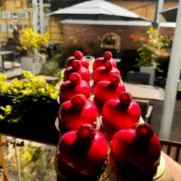 Rząd błyszczących, czerwonych deserów w kształcie serca z maliną na wierzchu, ułożonych na drewnianej desce, z widokiem na ogród w tle.