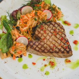 RAFAŁ KOZIORZEMSKI - Grillowany stek z tuńczyka z sałatką z marchewki, rzodkiewki i zieleniną, udekorowany sosem ziołowym i płatkami chili na białym talerzu z brązową obwódką.