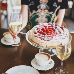 RAFAŁ KOZIORZEMSKI - Pavlova z truskawkami oprószona cukrem pudrem na szklanej paterze, dwie filiżanki cappuccino i kieliszki z białym winem na ciemnym stole, osoba w tle trzyma kieliszek w dłoni.