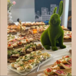 Stół z przekąskami i dekoracją w postaci zielonego zająca z mchu, jajka faszerowane na pierwszym planie, kanapki i tartaletki w tle, grafika zarysów miasta na ścianie.