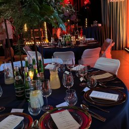 RAFAŁ KOZIORZEMSKI - Uroczysty stół z ciemnoniebieskim obrusem, nakryty złotymi sztućcami i talerzami z czerwonym obramowaniem, udekorowany wysokimi kompozycjami kwiatowymi i oświetlony ciepłym światłem, w tle scena...