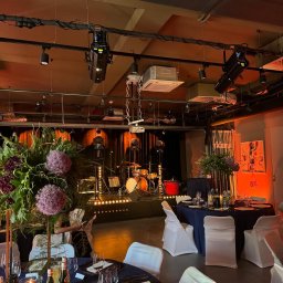 RAFAŁ KOZIORZEMSKI - Uroczysta sala z ustawionymi stołami z ciemnymi obrusami i białymi krzesłami, scena z oświetleniem i perkusją w tle, dekoracje kwiatowe z kulistymi kwiatami czosnku ozdobnego.