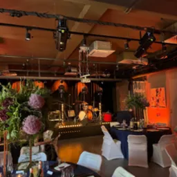 Uroczysta sala z ustawionymi stołami z ciemnymi obrusami i białymi krzesłami, scena z oświetleniem i perkusją w tle, dekoracje kwiatowe z kulistymi kwiatami czosnku ozdobnego.