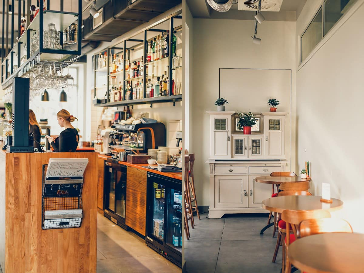 Wnętrze kawiarni z drewnianym barem, ekspres do kawy, półki z alkoholem i stoliki z krzesłami w tle, po prawej stronie kredens z roślinami doniczkowymi.