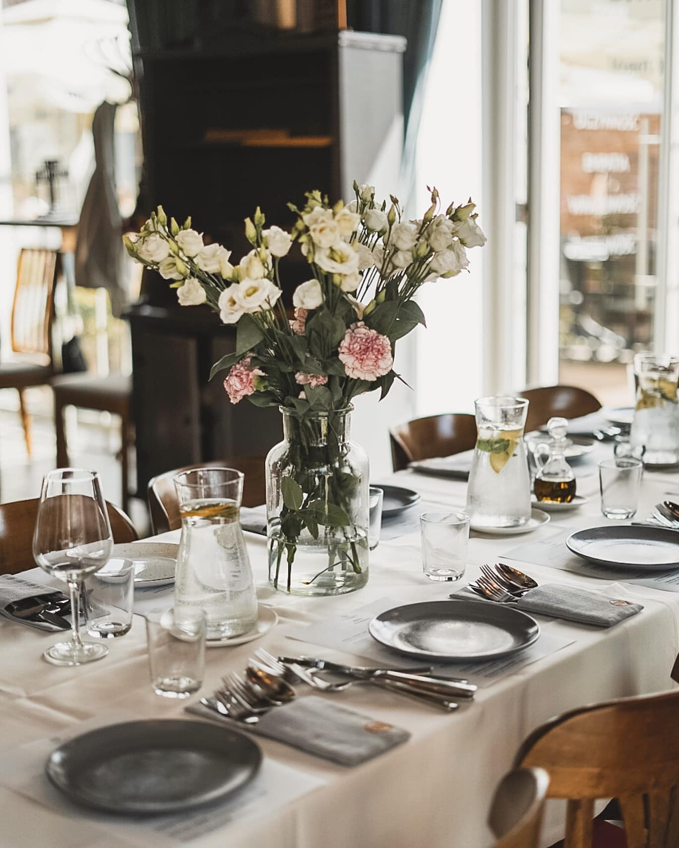 Elegancko nakryty stół z ciemnymi talerzami, srebrnymi sztućcami w szarych serwetach, szklanymi karafkami z wodą i cytryną oraz wazonem z białymi i różowymi kwiatami, ustawiony w jasnym...