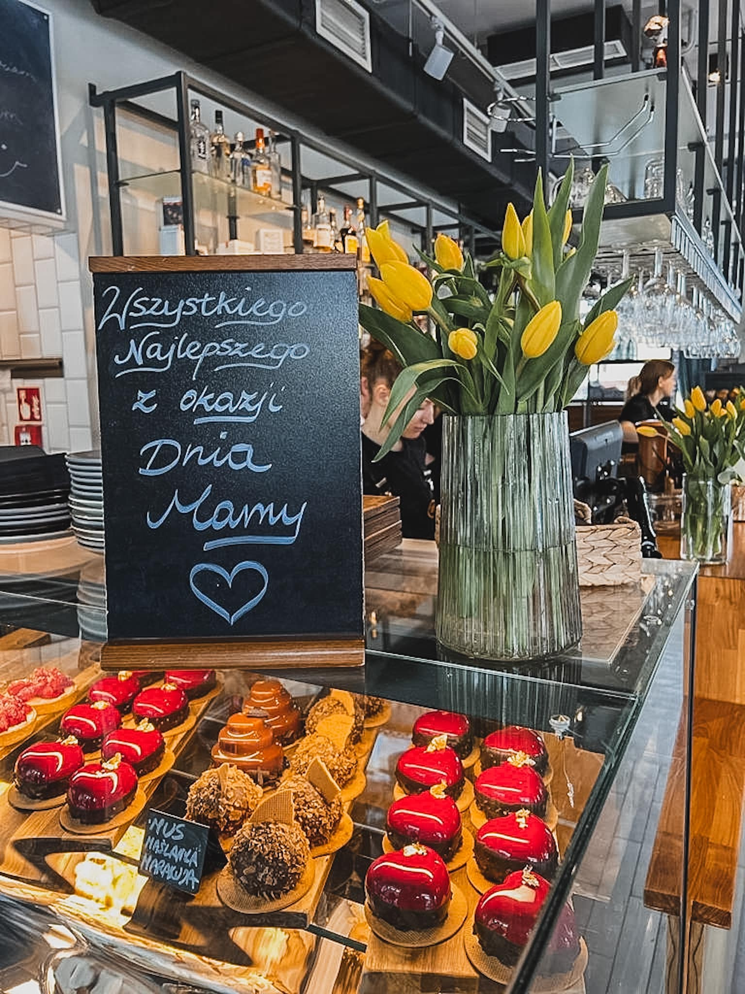 Witryna cukiernicza z różnorodnymi ciastkami i tortami, tablica z napisem 'Wszystkiego Najlepszego z okazji Dnia Mamy' i bukiet żółtych tulipanów w szklanym wazonie, widoczne elementy wystroju...