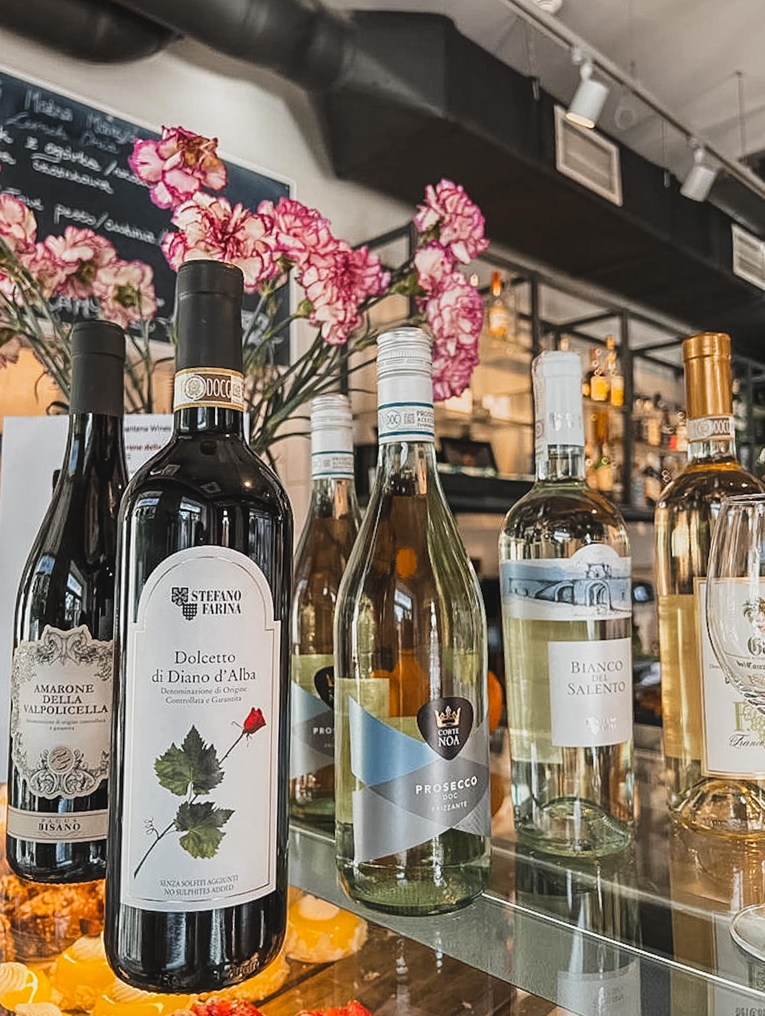 Różne butelki wina, w tym czerwone i białe, ustawione na szklanej półce z deserami w tle, ozdobione różowymi goździkami i tablicą z menu.