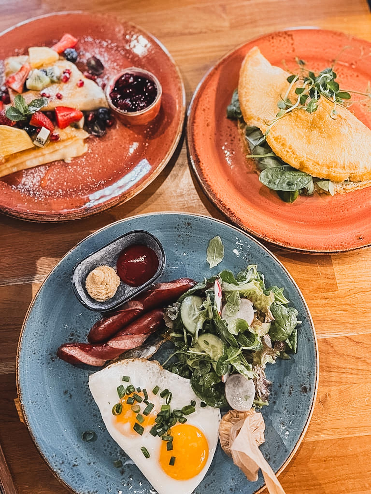 Kompozycja trzech talerzy z różnorodnymi daniami: naleśniki z owocami i dżemem, omlet ze szpinakiem i kiełkami oraz jajko sadzone z kiełbasą i sałatką, serwowana w Gdańsku.
