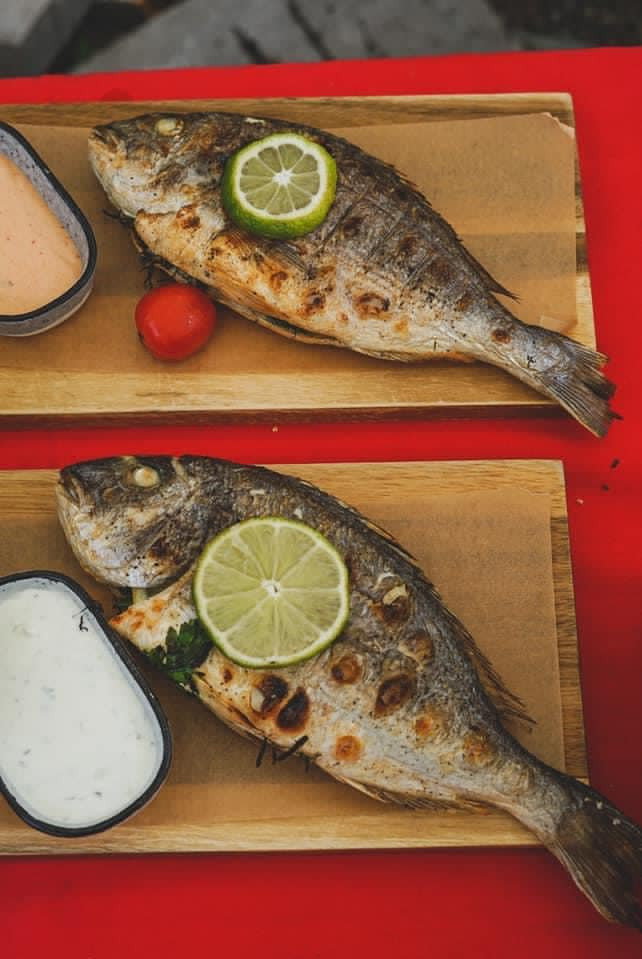 Dwie grillowane ryby dorady, każda podana na drewnianej desce z plasterkiem limonki i sosem w ceramicznej miseczce, ułożone na czerwonym obrusie.