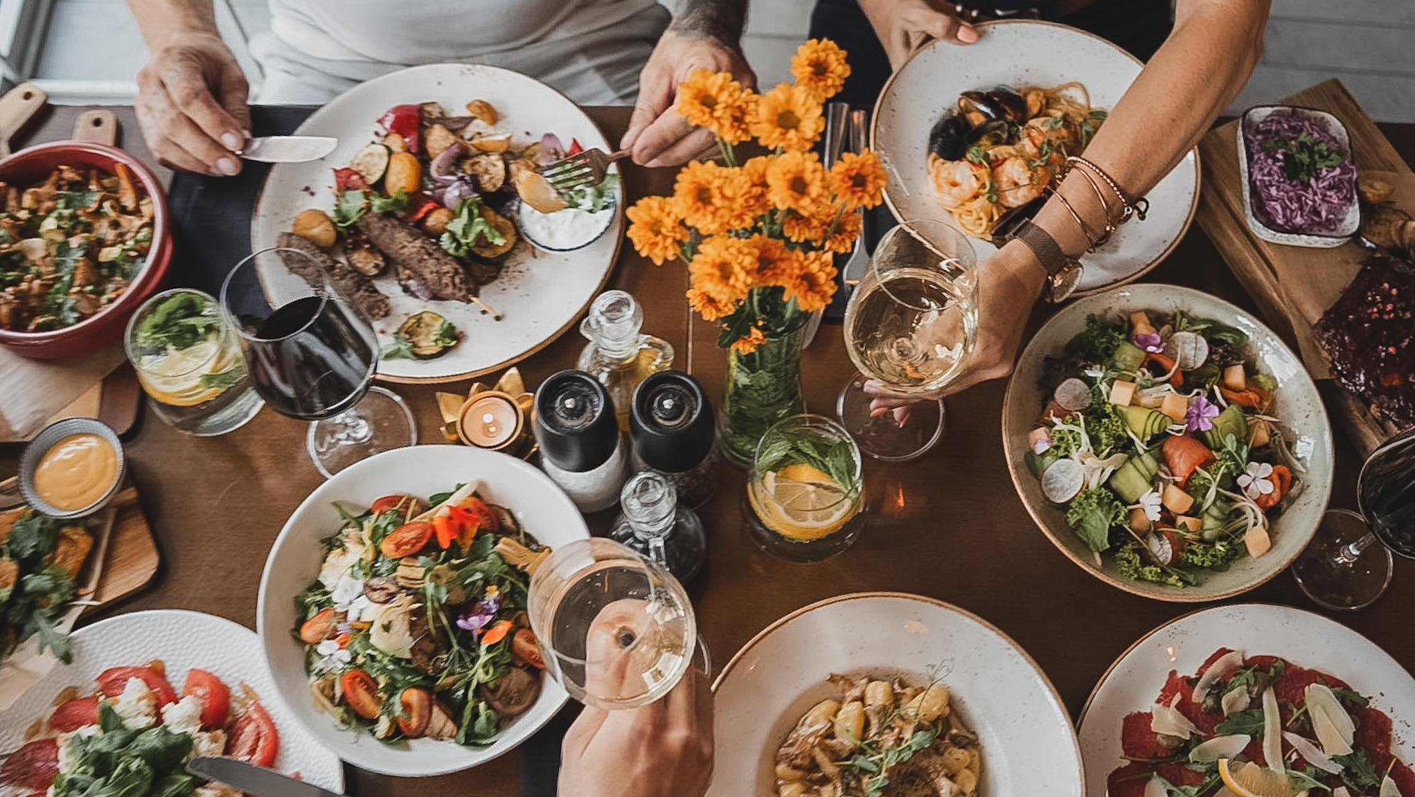 Stół z różnorodnymi daniami: sałatki z kwiatami, mięso z warzywami, owoce morza z makaronem, żeberka, napoje z cytryną, w tle osoby trzymające kieliszki z winem i sztućce.