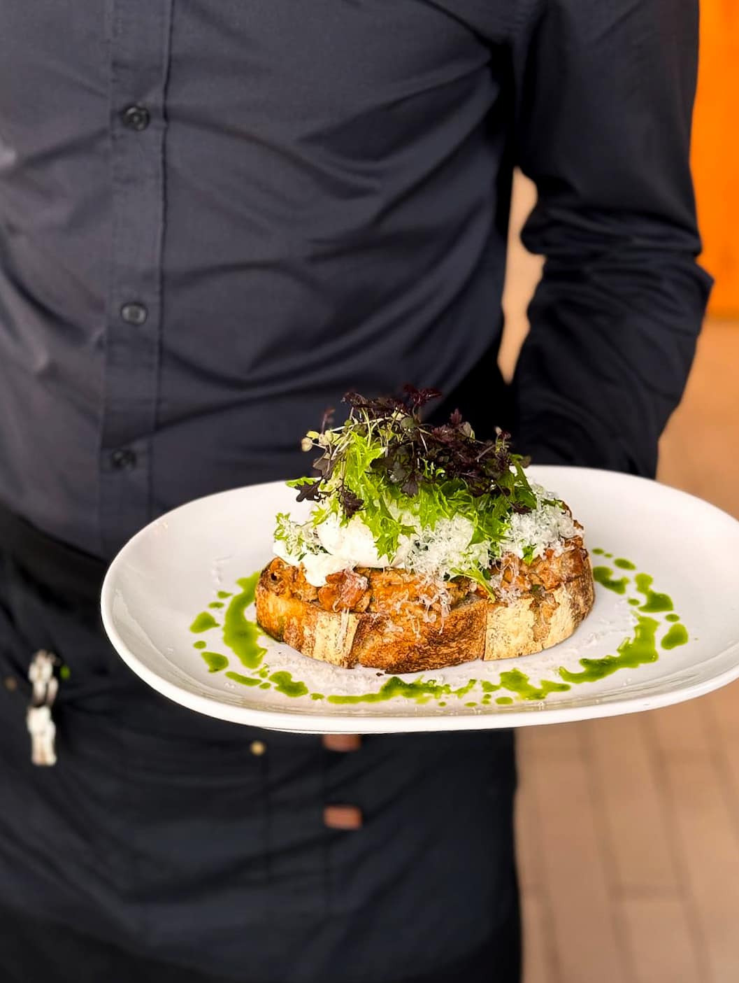 Elegancko podane danie na kromce chleba z dodatkiem zieleniny i sosem, trzymane przez kelnera w czarnym uniformie.