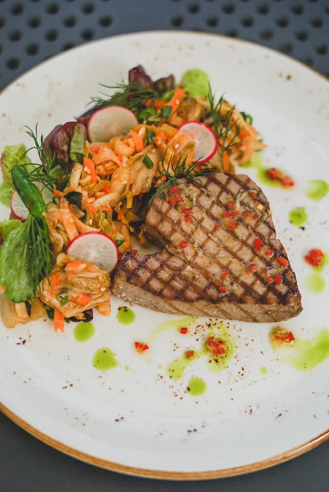 Grillowany stek z tuńczyka z sałatką z marchewki, rzodkiewki i zieleniną, udekorowany sosem ziołowym i płatkami chili na białym talerzu z brązową obwódką.