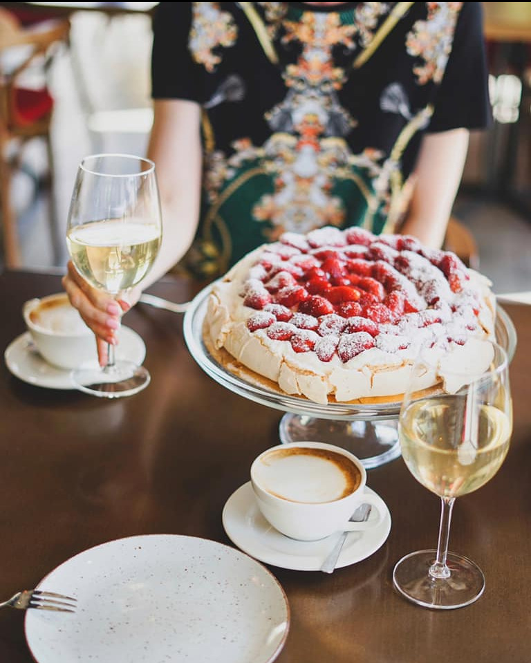 Pavlova z truskawkami oprószona cukrem pudrem na szklanej paterze, dwie filiżanki cappuccino i kieliszki z białym winem na ciemnym stole, osoba w tle trzyma kieliszek w dłoni.