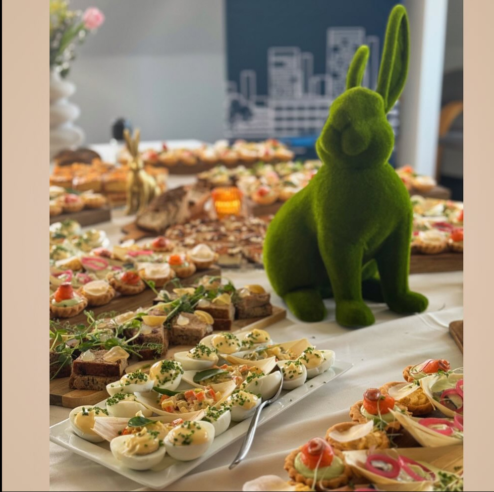 Stół z przekąskami i dekoracją w postaci zielonego zająca z mchu, jajka faszerowane na pierwszym planie, kanapki i tartaletki w tle, grafika zarysów miasta na ścianie.