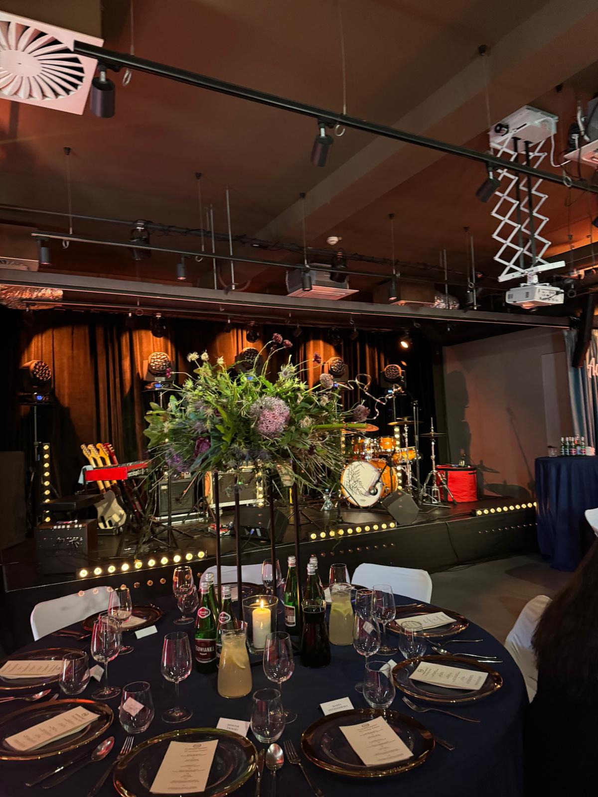 Uroczysta kolacja na scenie z instrumentami muzycznymi w tle, dekoracja kwiatowa na stole, elegancka zastawa, napoje w szklanych dzbankach i butelkach.