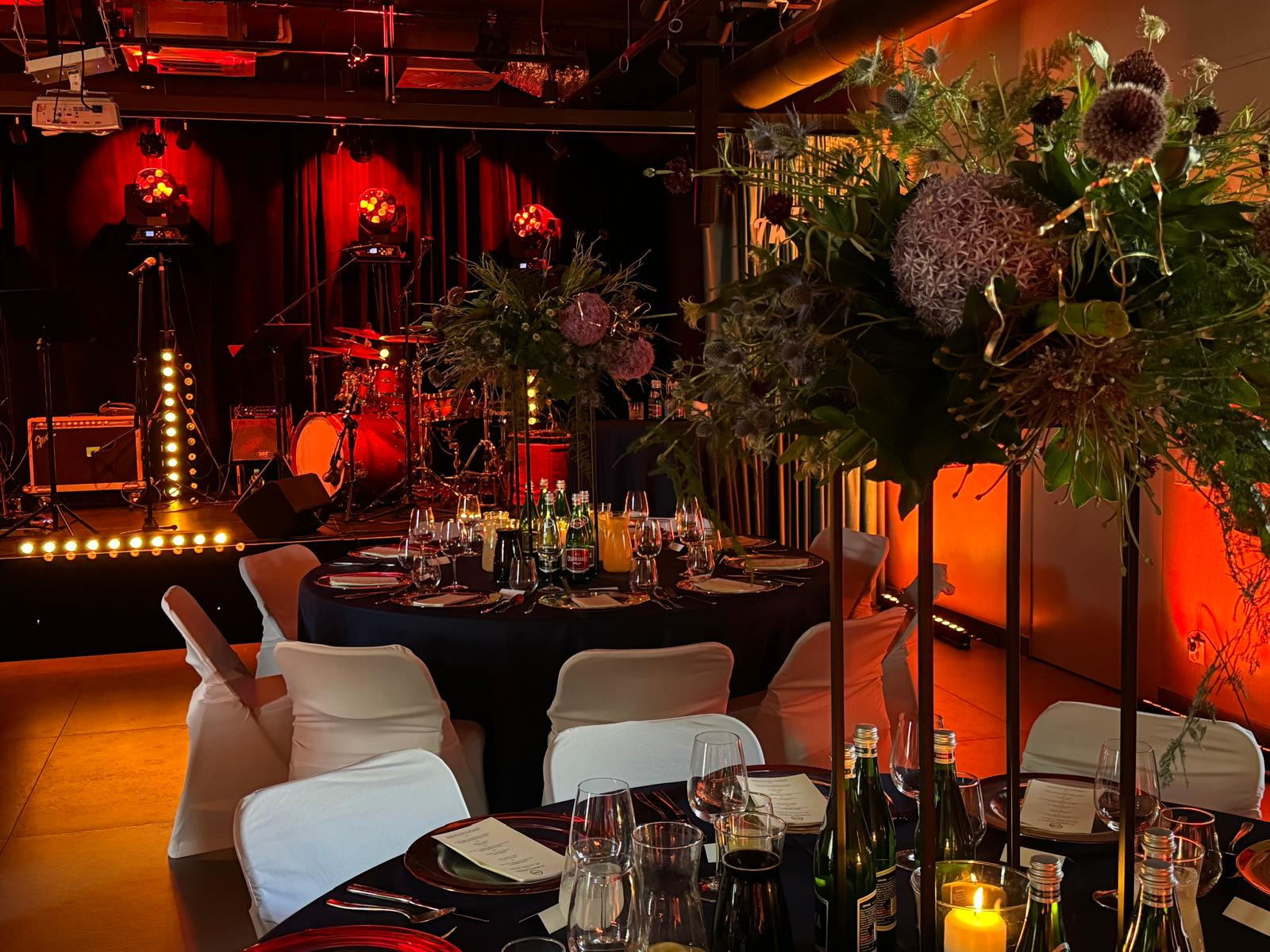 Uroczysta sala z okrągłymi stołami nakrytymi ciemnymi obrusami, białymi pokrowcami na krzesłach i wysokimi kompozycjami kwiatowymi, w tle scena z instrumentami oświetlona czerwonymi światłami.