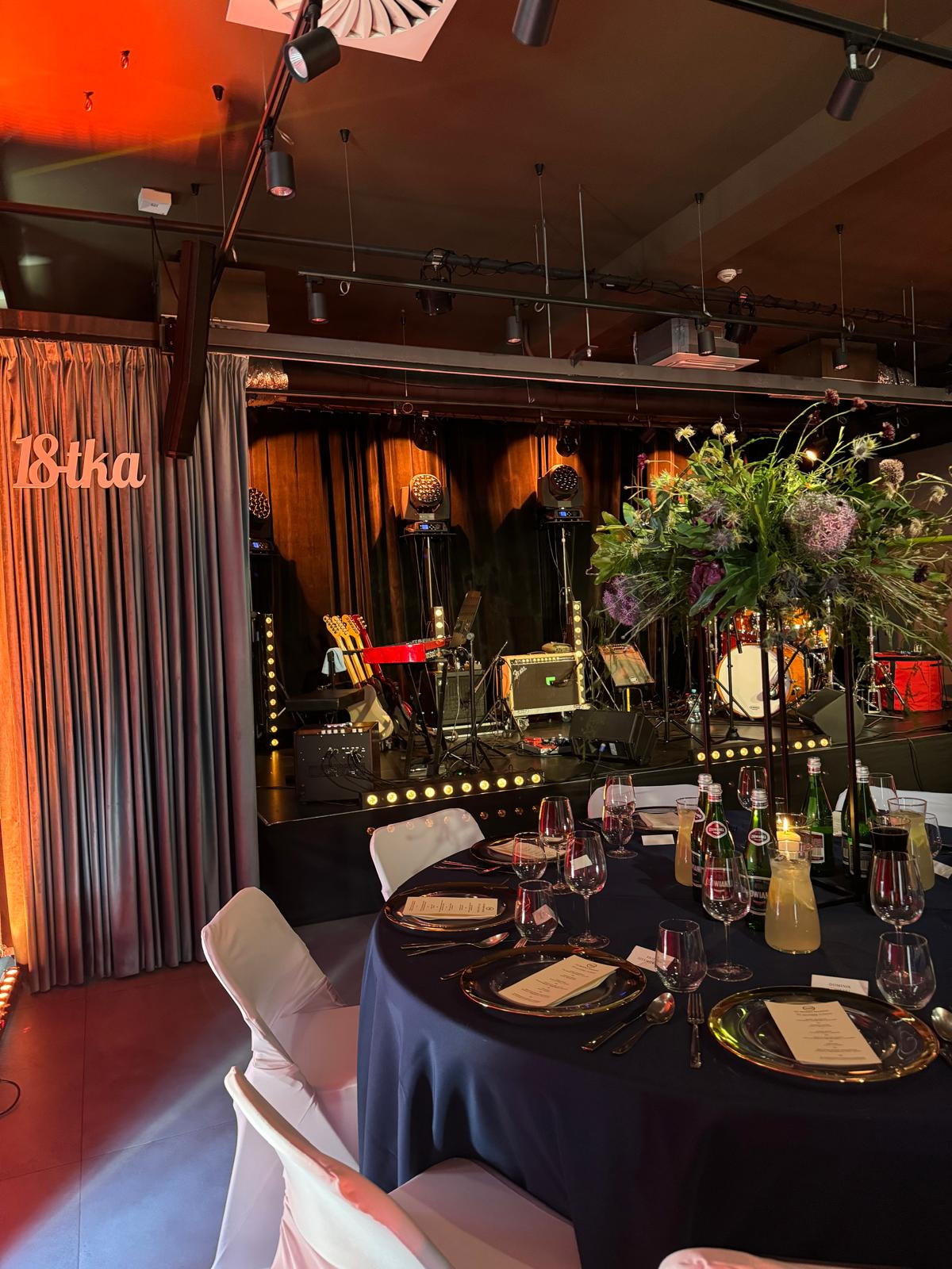 Uroczysta sala z przygotowanym stołem na osiemnastkę, scena z instrumentami muzycznymi w tle, oświetlenie punktowe, dekoracja kwiatowa na stole.