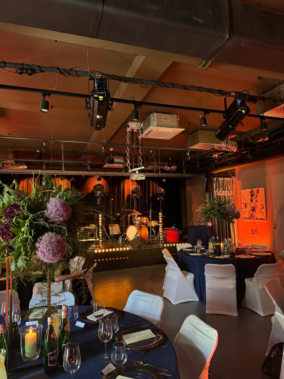 Uroczysta sala z ustawionymi stołami z ciemnymi obrusami i białymi krzesłami, scena z oświetleniem i perkusją w tle, dekoracje kwiatowe z kulistymi kwiatami czosnku ozdobnego.