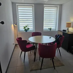 Jasne pomieszczenie z okrągłym stołem, czterema bordowymi krzesłami, lampą podłogową i komodą. Dwa okna z żaluzjami w tle.